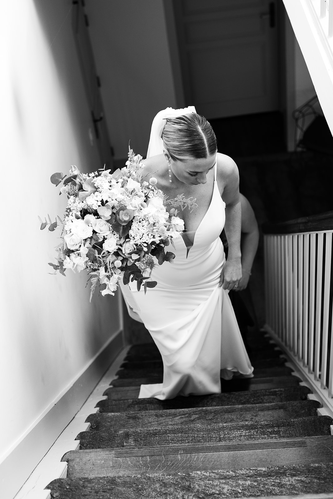 A bride in a white dress, adorned with a veil and a bouquet, walks down a staircase in a black-and-white photograph.